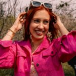 Smiling woman in a vibrant pink jacket enjoying a sunny day outdoors with stylish accessories.